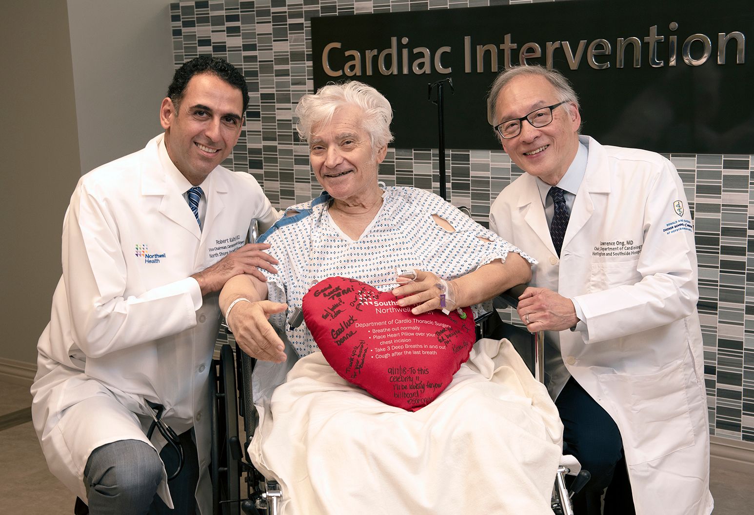 An elderly male patient with white hair wearing a patient gown is sitting in a wheel chair holding a heart pillow. Two doctors wearing Northwell branded white coats kneel beside him and smile.
